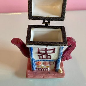 May include: A small ceramic teapot trinket box with a hinged lid. The teapot is painted to look like a house with a red roof, blue trim, and white walls. The teapot has a pink base and a red handle.