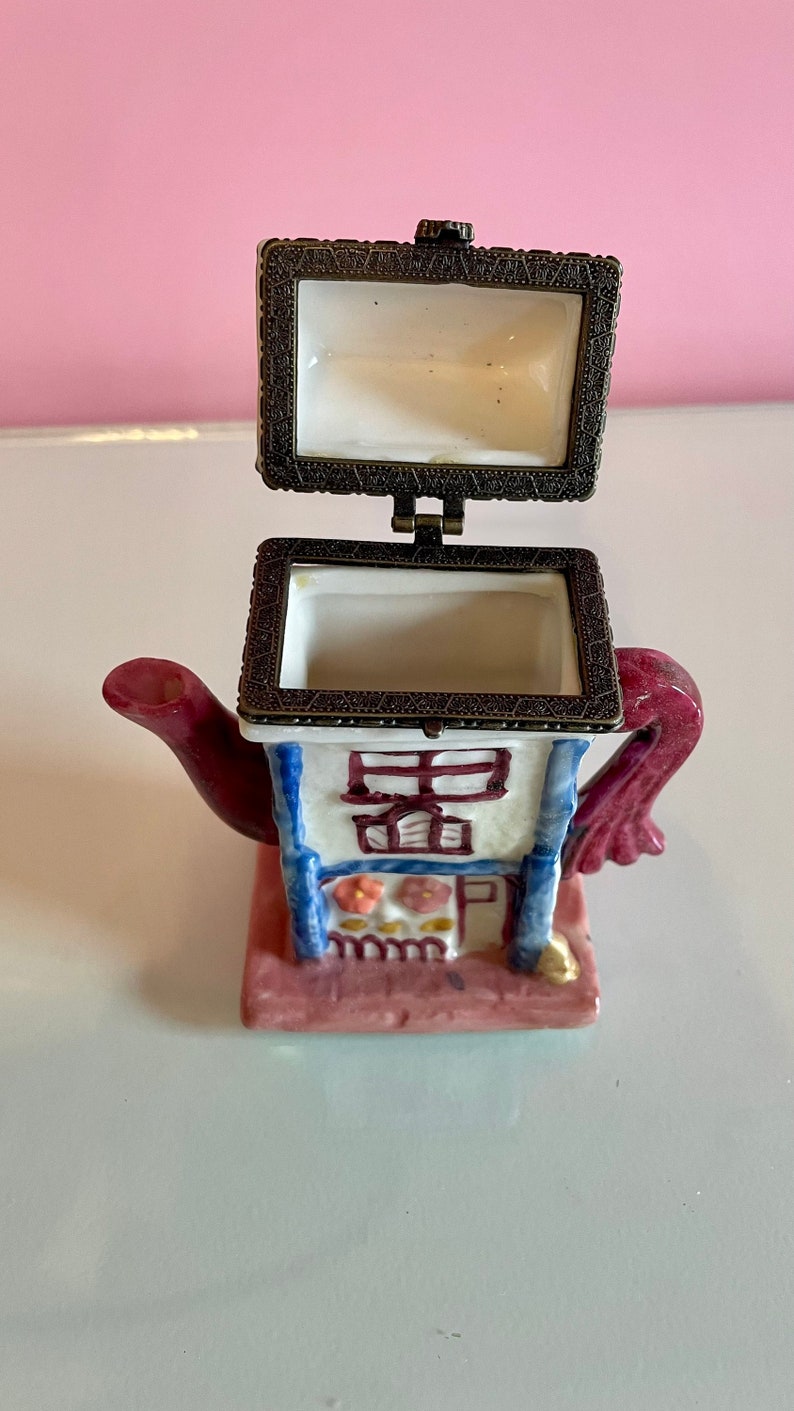 May include: A small ceramic teapot trinket box with a hinged lid. The teapot is painted to look like a house with a red roof, blue trim, and white walls. The teapot has a pink base and a red handle.