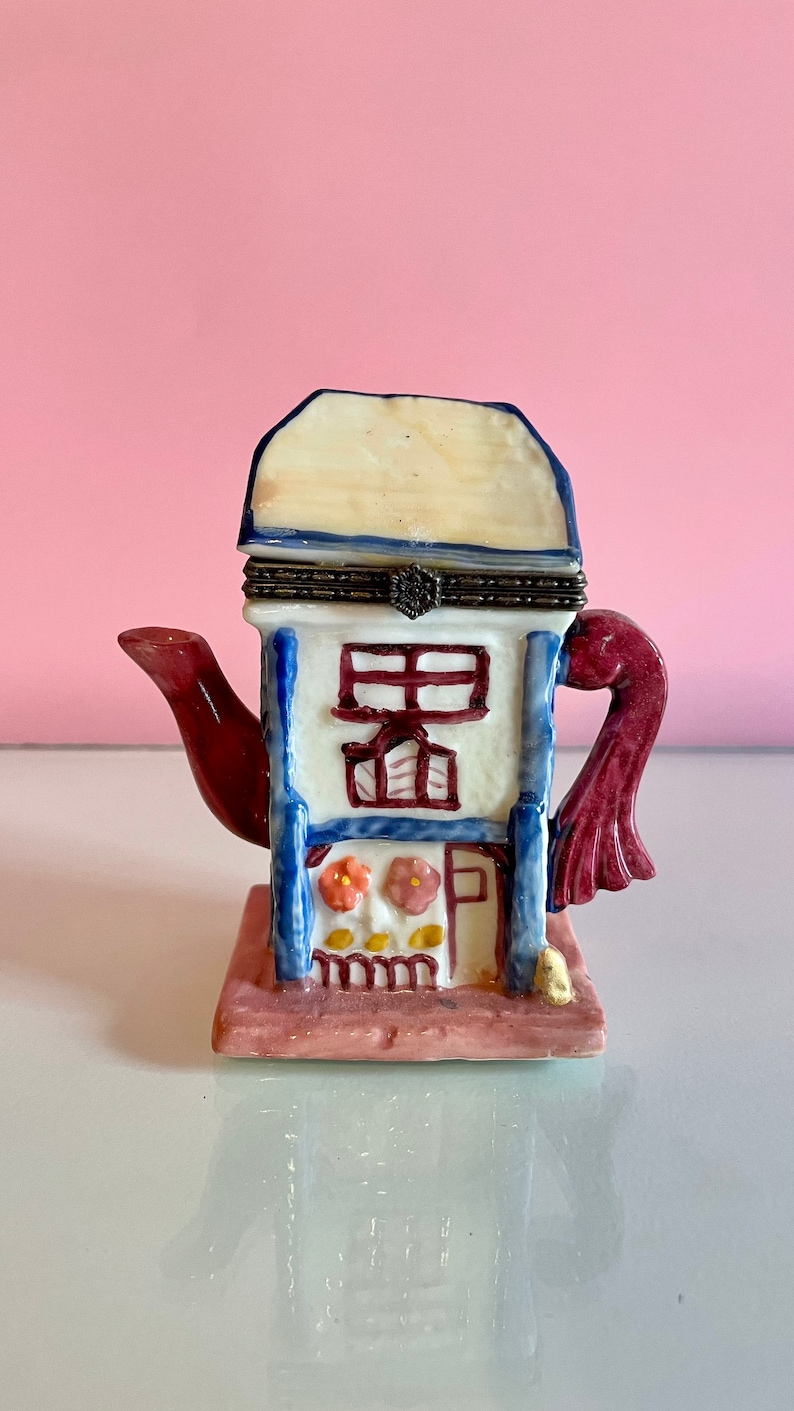 May include: A ceramic teapot shaped like a house with a red spout and a burgundy handle. The house has a white base with blue trim and a red roof. The front of the house has a window and a door with a floral design. The house is sitting on a pink base.