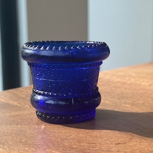 Pode incluir: Um tampão de garrafa de vidro azul cobalto com um design decorativo. O tampão tem uma parte superior e inferior arredondadas, com um corpo texturizado e padronizado. A peça tem aproximadamente 5 cm de altura.