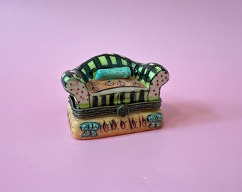 May include: A small ceramic trinket box shaped like a couch with a black and green striped pattern, red polka dots, and a light blue pillow. The base of the box is yellow with a green and blue floral design.