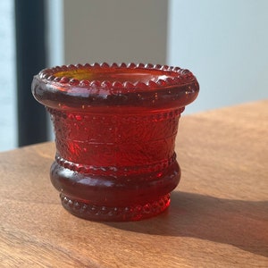 Pode incluir: Um pequeno vaso de vidro vermelho com um padrão decorativo. O vaso tem uma borda com contas e uma base arredondada.
