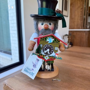 Könnte beinhalten: Eine Holz-Nussknacker-Figur mit schwarzem Zylinder, grünem Band und Kunstpelz-Bart. Sie hat ein Kuckucksuhr-Design auf der Brust und ein Etikett mit der Aufschrift "Christian Ulbricht". Ein Schlüssel liegt auf der Holzoberfläche.