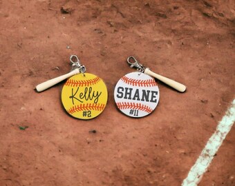 May include: Two personalized softball keychains with a wooden bat charm. One keychain is yellow with the name "Kelly" and the number "#2". The other keychain is white with the name "Shane" and the number "#11".