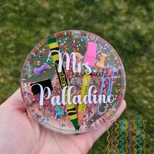 May include: A clear, round coaster with a colorful confetti design. The coaster features a yellow crayon, a pencil, push pins, paper clips, and the name "Mrs. Palladino" in white script. The coaster is a teacher-themed craft.
