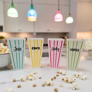 Pode incluir: Quatro recipientes de papel para pipocas com bordas superiores recortadas, apresentando listras verticais em turquesa, amarelo, rosa e verde claro. Cada recipiente exibe uma etiqueta oval central com os nomes Jess, Clark, Haley e Harper.