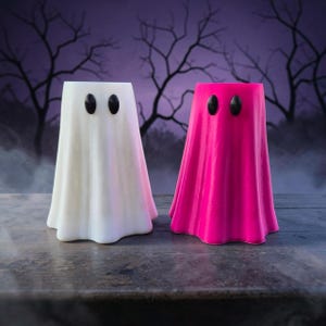 May include: Two ghost-shaped decorative figures, one in white and one in bright pink, each featuring a smooth, draped silhouette and two small, oval-shaped black eyes positioned near the top. The figures stand upright with a hollow, open top.