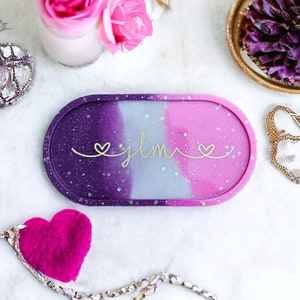May include: A purple, white, and pink oval tray with glitter embedded in the resin. The tray has a gold inscription that reads "jlmi" with a heart on each side of the name.