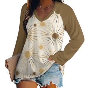 Op de afbeelding: Een shirt met lange mouwen in een bruin en wit kleurenschema. Het shirt heeft een V-hals en een bruin raglan mouwontwerp. De voorkant van het shirt heeft een witte achtergrond met een patroon van gouden en zwarte gestileerde sterren.