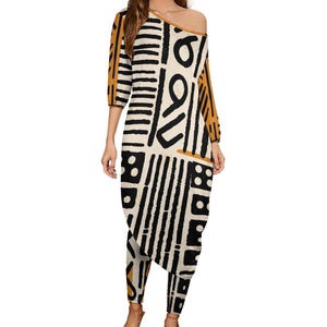 Op de afbeelding: Een witte, zwarte en mosterdgele geprinte jurk met een lange, asymmetrische zoom. De jurk heeft driekwartmouwen en een off-shoulder halslijn.