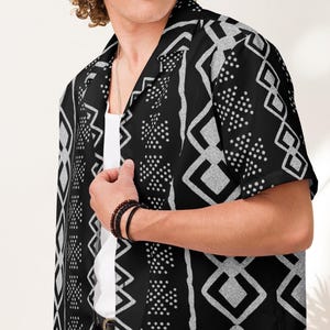 Puede incluir: Camisa negra de manga corta con un estampado geométrico blanco. La camisa tiene cuello de campamento y se usa sobre una camiseta interior blanca. El diseño presenta diamantes, puntos y líneas verticales.