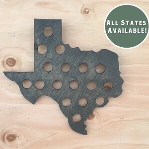 Puede incluir: Un recorte de madera del estado de Texas con una mancha marrón oscura. El recorte tiene 30 agujeros para tapas de botellas. El texto "ALL STATES AVAILABLE!" está escrito en blanco sobre un círculo verde.