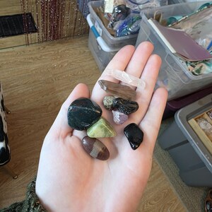 Könnte beinhalten: Eine Hand hält eine Sammlung polierter Edelsteine und Kristalle. Die Steine sind in verschiedenen Farben, darunter Schwarz, Grün, Braun und Klar. Die Sammlung umfasst eine Vielzahl von Formen und Größen, einige glatt und andere strukturiert.