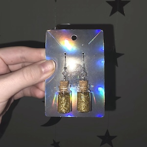 May include: A pair of earrings featuring small glass bottles filled with gold glitter, each bottle is sealed with a cork stopper. The earrings hang from silver-colored hooks and are displayed on a holographic card.