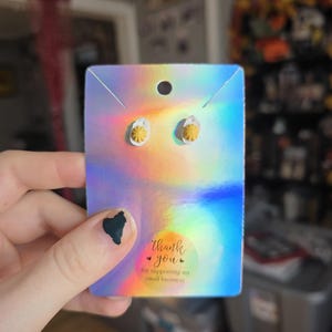 May include: A pair of small, yellow flower stud earrings on a holographic card. The card has a thank you message for supporting a small business. The earrings are held by a hand with a dark green nail design.