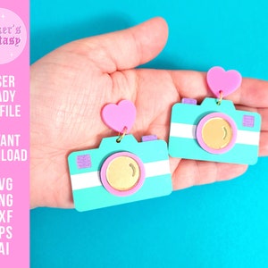 Puede incluir: Un par de pendientes de cámara azul turquesa con corazones rosas y detalles dorados. Los pendientes están cortados con láser y listos para ser hechos.