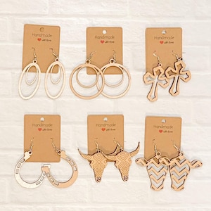 May include: A collection of handmade wooden earrings in various shapes, including oval, hoop, cross, horseshoe, bull skull, and cow designs. Each pair is attached to a brown card with the text "Handmade with love".