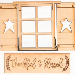 Op de afbeelding: Een houten bord met een raamkozijn en luiken. Het bord heeft de tekst "thankful & blessed" in een decoratief lettertype.