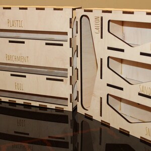 May include: A wooden organizer with labeled compartments for storing different types of food wrap. The compartments are labeled "Plastic", "Parchment", "Foil", "Snack", "Sandwich", "Quart", and "Gallon".