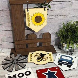 Puede incluir: Un poste de madera con una variedad de letreros de madera colgantes. Los letreros presentan un girasol, una abeja, un automóvil, una vaca y una estrella. Los letreros están pintados en varios colores, incluyendo amarillo, azul, rojo, blanco y negro.