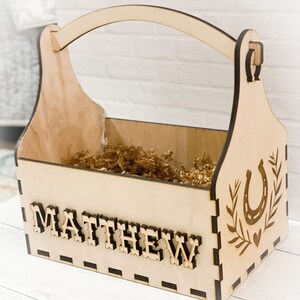 Puede incluir: Una caja de madera con asa, personalizada con el nombre "MATTHEW". La caja tiene un diseño de herradura y está llena de papel de embalaje marrón.