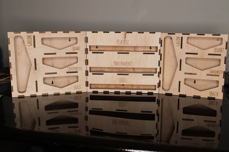 May include: Wooden organizer with slots for storing various sizes of plastic wrap, parchment paper, and aluminum foil. The organizer has labels for "Gallon", "Quart", "Sandwich", and "Snack".