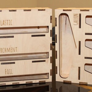 May include: A wooden storage box with two sections. The left section has three slots labeled "Plastic", "Parchment", and "Foil". The right section has four slots labeled "Gallon", "Quart", "Sandwich", and "Snack".