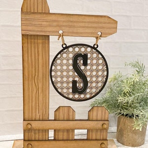 Puede incluir: Un letrero de madera con un diseño de valla de piquetes y un letrero redondo colgante con una letra S negra sobre un patrón tejido. El letrero está sobre una base de madera.
