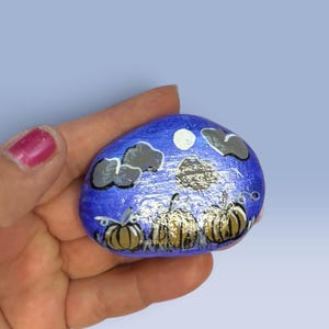 May include: A smooth, oval-shaped river rock painted with a nighttime scene featuring three golden-brown pumpkins in the foreground. The background is a vibrant blue sky with a white circular moon and several dark gray clouds painted above the pumpkins.
