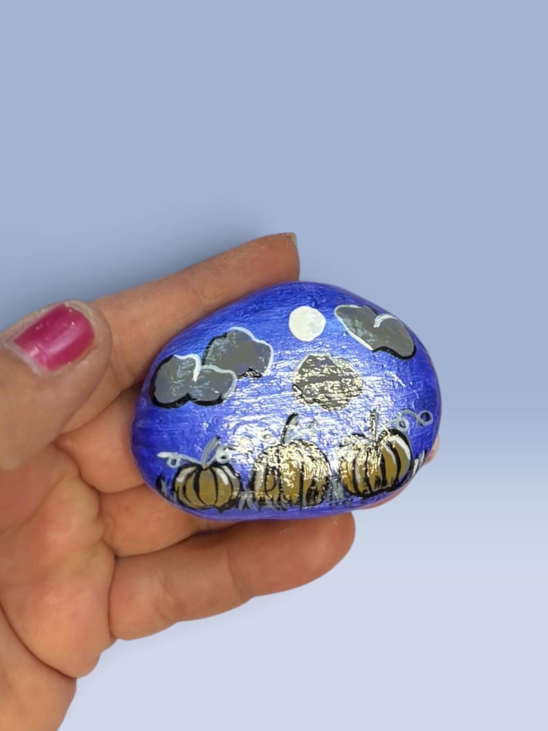 May include: A smooth, oval-shaped river rock painted with a nighttime scene featuring three golden-brown pumpkins in the foreground. The background is a vibrant blue sky with a white circular moon and several dark gray clouds painted above the pumpkins.