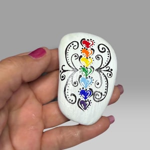 May include: A smooth, white, oval-shaped stone featuring a vertical column of seven small, hand-painted hearts in rainbow colors: red, orange, yellow, green, light blue, dark blue, and purple, surrounded by intricate black swirling line art.