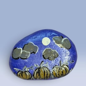 May include: A smooth, oval-shaped river rock painted with a nighttime scene featuring three golden-brown pumpkins in the foreground. Above the pumpkins, the dark blue sky displays a pale yellow full moon and several dark grey, fluffy clouds.