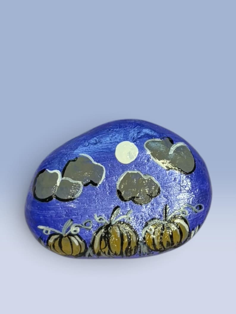 May include: A smooth, oval-shaped river rock painted with a nighttime scene featuring three golden-brown pumpkins in the foreground. Above the pumpkins, the dark blue sky displays a pale yellow full moon and several dark grey, fluffy clouds.