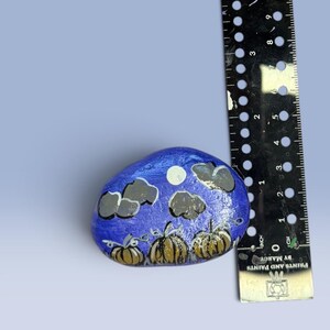 May include: A hand-painted rock featuring a night scene with three brown pumpkins, a white moon, and grey clouds against a blue background, positioned next to a black ruler showing a width of approximately 2.5 inches for the painted stone.