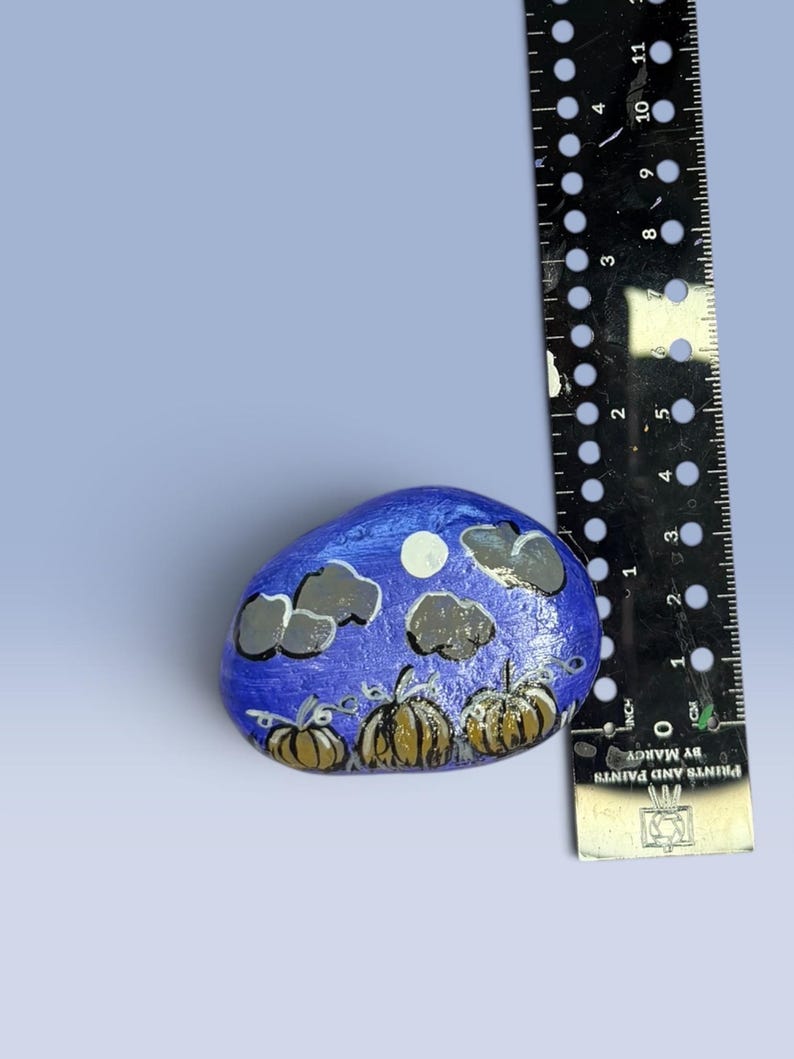 May include: A hand-painted rock featuring a night scene with three brown pumpkins, a white moon, and grey clouds against a blue background, positioned next to a black ruler showing a width of approximately 2.5 inches for the painted stone.