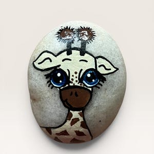 May include: A smooth, oval-shaped light gray stone hand-painted with a cartoon giraffe face. The giraffe features large, bright blue eyes with long black eyelashes, a brown muzzle, and two small brown ossicones with dark brown tufts on top.