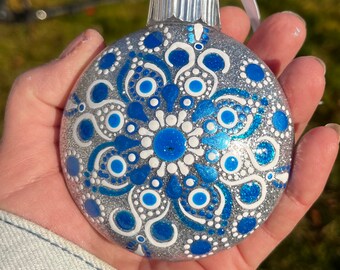 May include: Silver glitter Christmas ornament with a blue and white mandala design. The ornament has a silver cap and a white ribbon for hanging. The mandala features intricate patterns and a central flower design.