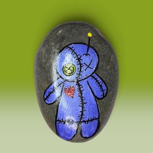 May include: A smooth, oval-shaped gray river rock featuring a hand-painted blue voodoo doll character. The doll has a lime green button eye, a red heart patch on its chest, black stitched details, and a single yellow pin sticking out of its head.
