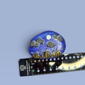 May include: A hand-painted rock featuring a night scene with three orange pumpkins, dark grey clouds, and a white moon against a blue background. A black ruler is positioned underneath, showing the rock is approximately 2.5 inches wide.