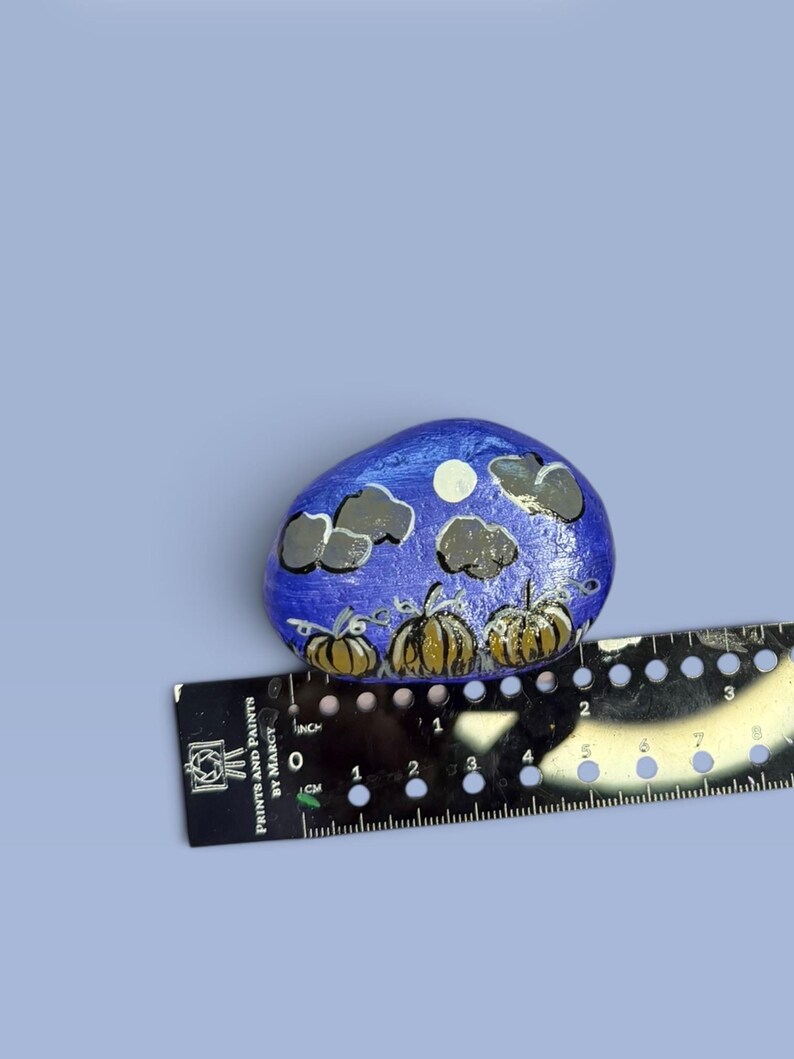 May include: A hand-painted rock featuring a night scene with three orange pumpkins, dark grey clouds, and a white moon against a blue background. A black ruler is positioned underneath, showing the rock is approximately 2.5 inches wide.