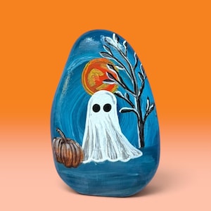 May include: A smooth, oval-shaped river rock painted with a nighttime scene featuring a white ghost with two black circular eyes, a brown pumpkin, a bare black tree, and a bright orange moon against a dark blue, swirled sky background.