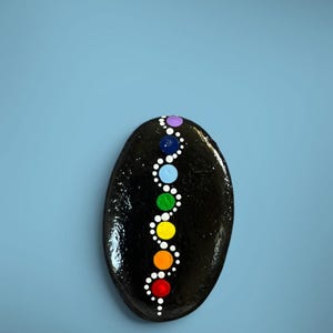 May include: A smooth, oval-shaped black stone featuring a vertical, winding pattern of painted dots. The design includes seven larger colored dots in purple, dark blue, light blue, green, yellow, orange, and red, surrounded by smaller white dots.
