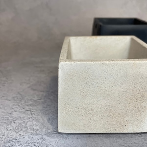 Peut inclure: Un pot en béton blanc de forme carrée et de texture rugueuse. Le pot est vide et prêt à être rempli de plantes.