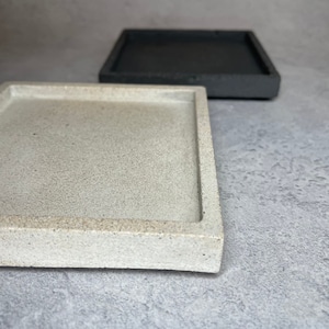 Peut inclure: Deux plateaux carrés en béton, l'un de couleur beige clair et l'autre de couleur gris foncé. Les plateaux sont tous deux faits d'un matériau rugueux et texturé.