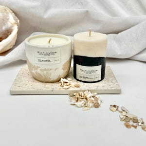 May include: Two concrete candles with black and white designs. The candles are on a white and brown speckled surface. The candles are labelled "Marmalade Lane" and "OUD & NEROLI" with the number "2" on the label. There are white and brown pieces of dried flowers scattered around the candles.