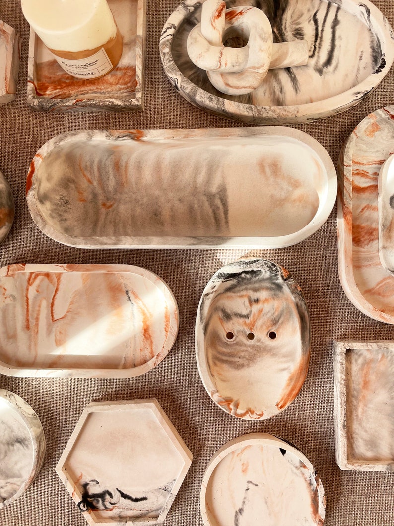 Pu&ograve; includere: Una collezione di vassoi decorativi in ​​calcestruzzo con un design marmoreo in tonalit&agrave; di bianco, grigio e arancione. I vassoi sono di varie forme e dimensioni, tra cui ovali, rotondi, quadrati ed esagonali.