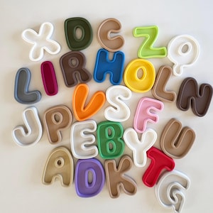 Puede incluir: Un conjunto de letras del alfabeto coloridas en varios tonos, incluyendo blanco, verde, marrón y rosa. Las letras están dispuestas sobre un fondo claro, mostrando sus diversos colores y formas. Ideal para fines educativos o decorativos.