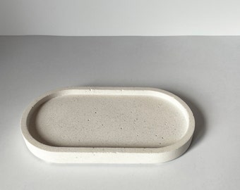 Puede incluir: Una bandeja ovalada de hormigón de color claro. La bandeja tiene una superficie lisa, ligeramente texturizada y bordes redondeados. El color neutro y el diseño minimalista la hacen adecuada para diversos usos, como pieza decorativa o para guardar objetos pequeños.