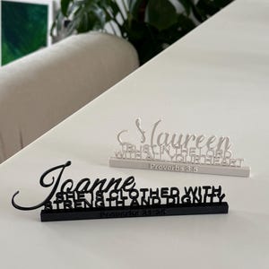 May include: Two custom name signs on rectangular bases. One white sign features the name Maureen with the text TRUST IN THE LORD WITH ALL YOUR HEART Proverbs 3:5. One black sign features the name Joanne with the text SHE IS CLOTHED WITH STRENGTH AND DIGNITY Proverbs 31:25.