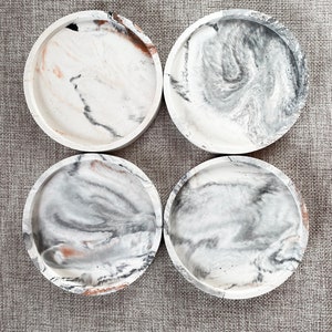 Pu&ograve; includere: Quattro sottobicchieri rotondi in cemento con un designo marmoreo in tonalit&agrave; di bianco, grigio e marrone.